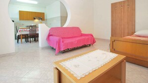 A room with a wooden bed, a pink sofa, a dining table with chairs, and a kitchen area visible through an archway. - Holiday apartment with air conditioning (Štinjan)