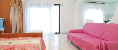 A hotel room with a bed, a pink sofa, a wooden wardrobe, a television, and a bathroom visible through a glass door.