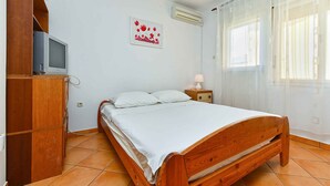 A bedroom with a wooden bed, a bedside table with a lamp, a TV on a wooden shelf, and a window with sheer curtains. - Studio with air conditioning (Pula)