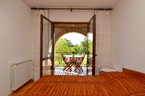 Une chambre avec un lit en bois, une vue sur un balcon avec une table et des chaises, accessible par une porte ouverte, et une arche en pierre.