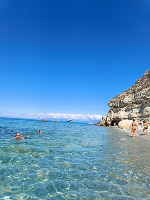 A beach with clear blue water, rocky cliffs, and people swimming and walking along the shore. - Comfortable Deluxe Apartment in the Center of Tropea with a Balcony Overlooking the Sea (Tropea)