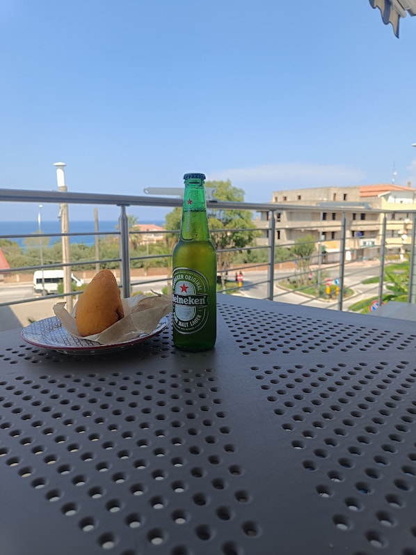 A green Heineken bottle and a cone-shaped snack on a perforated metal table with a view of buildings and a clear sky in the background. - Comfortable Deluxe Apartment in the Center of Tropea with a Balcony Overlooking the Sea (Tropea)