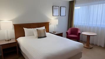 Una habitación de hotel con una cama grande, dos mesitas de noche con lámparas, una silla, una mesita redonda pequeña y una ventana con cortinas traslúcidas.