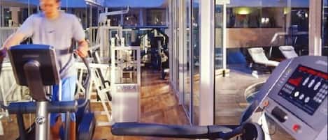A gym with cardio equipment and weight machines, large windows, and a wooden floor.