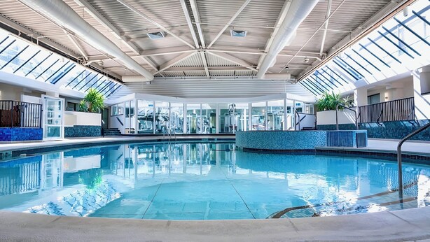 An indoor swimming pool with clear glass walls, a high ceiling with visible ductwork, and a tiled edge. - The Aberdeen Dyce Hotel (Aberdeen)