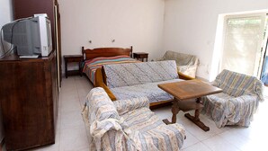 Habitació petita i ben il·luminada amb un llit, dues cadires, una taula petita i un televisor sobre un moble de fusta.