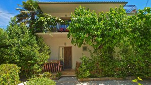 Edifici de dues plantes amb un balcó, envoltat de vegetació abundant i una zona de pati amb rajoles.