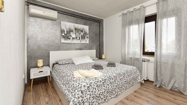 A modern bedroom with a large bed, a bedside table with a lamp, a wall-mounted air conditioner, and a window with curtains. - Holiday apartment with air conditioning (Rovinj)