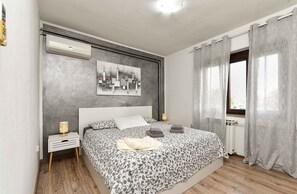 A modern bedroom with a large bed, a bedside table with a lamp, a wall-mounted air conditioner, and a window with curtains. - Holiday apartment with air conditioning (Rovinj)