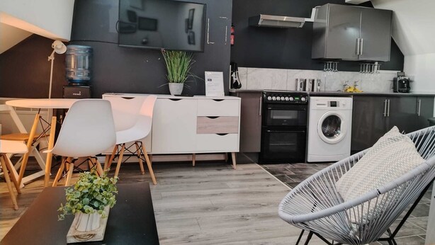 Une cuisine moderne équipée d’un four encastré, d’un four à micro-ondes et d’un lave-linge. On y trouve un coin repas avec une table et des chaises, ainsi qu’un coin salon avec un canapé et un fauteuil.