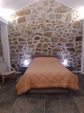 A bedroom with a stone wall, a bed with a quilt, a chair, and bedside lamps.