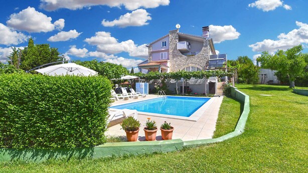 A two-story stone house with a swimming pool, surrounded by well-maintained lawns and potted plants.