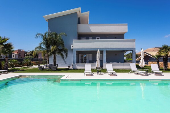A modern building with a pool, lounge chairs, and umbrellas. - Etna Mare Apartments by Wonderful Italy (Augusta)