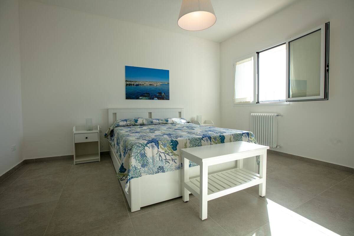 Ein Schlafzimmer mit Bett, Nachttischen, einem kleinen Tisch, einem Fenster und einer Heizung.