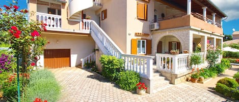 A two-story house with a balcony, a staircase, and a garden with potted plants.
