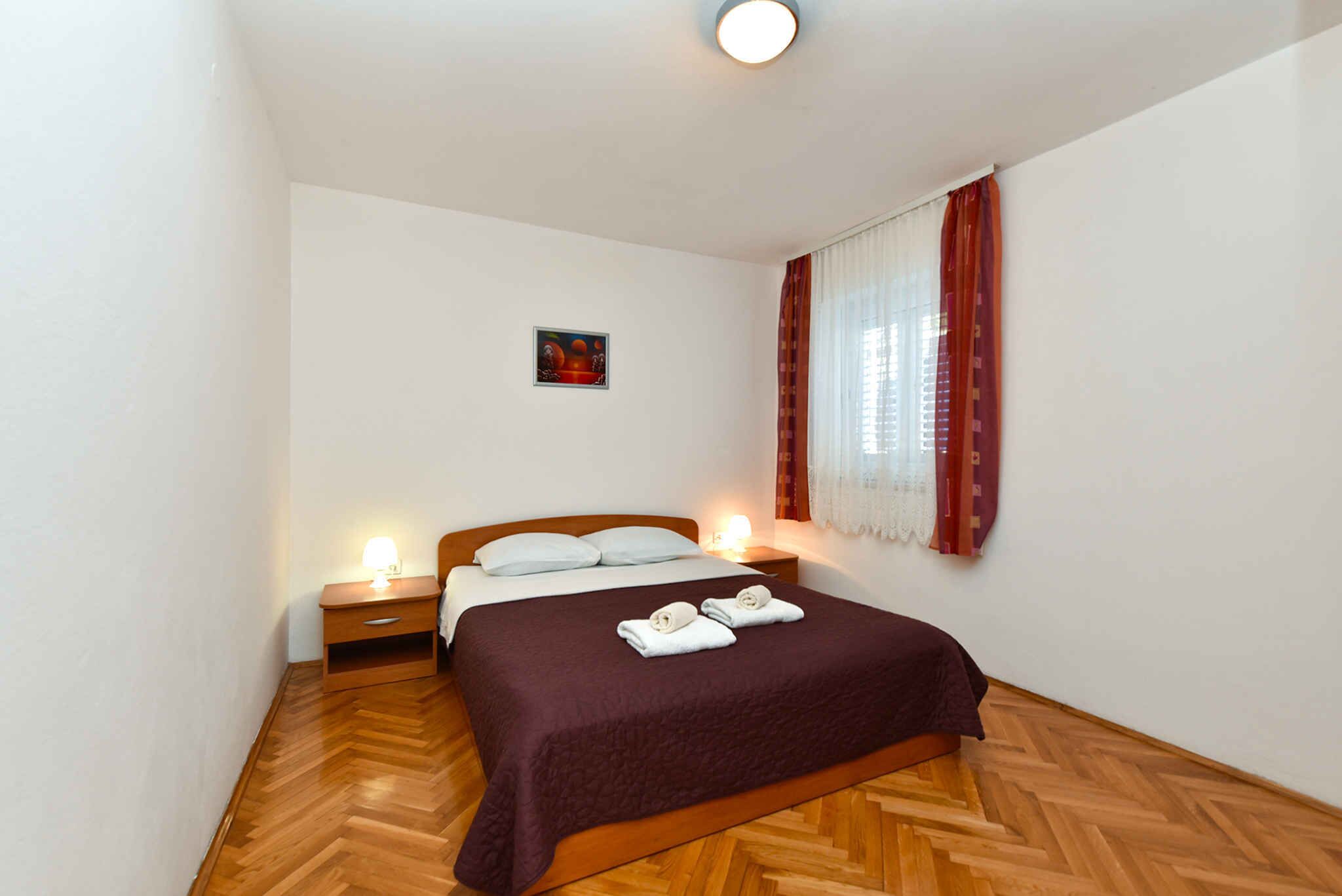 A bedroom with a wooden herringbone floor, a bed with a dark brown bedspread, two bedside lamps, and a window with red curtains.