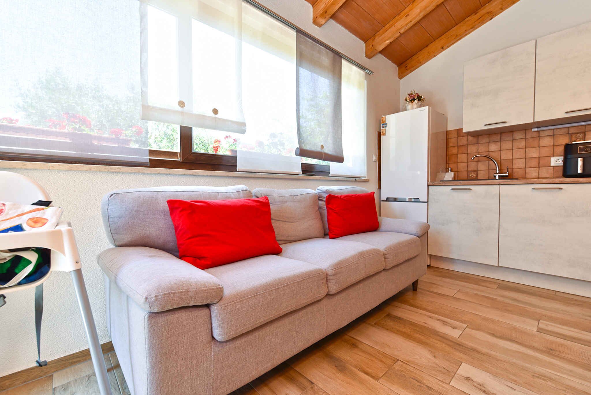 Eine moderne Küche mit Holzboden, einem grauen Sofa und roten Kissen sowie einem Fenster mit Jalousien.