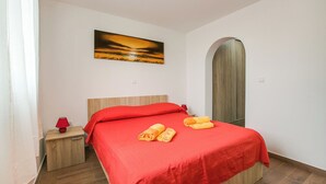 A neatly made bed with red bedding and two rolled towels, a small bedside table with a red lamp, and a painting of a sunset on the wall.