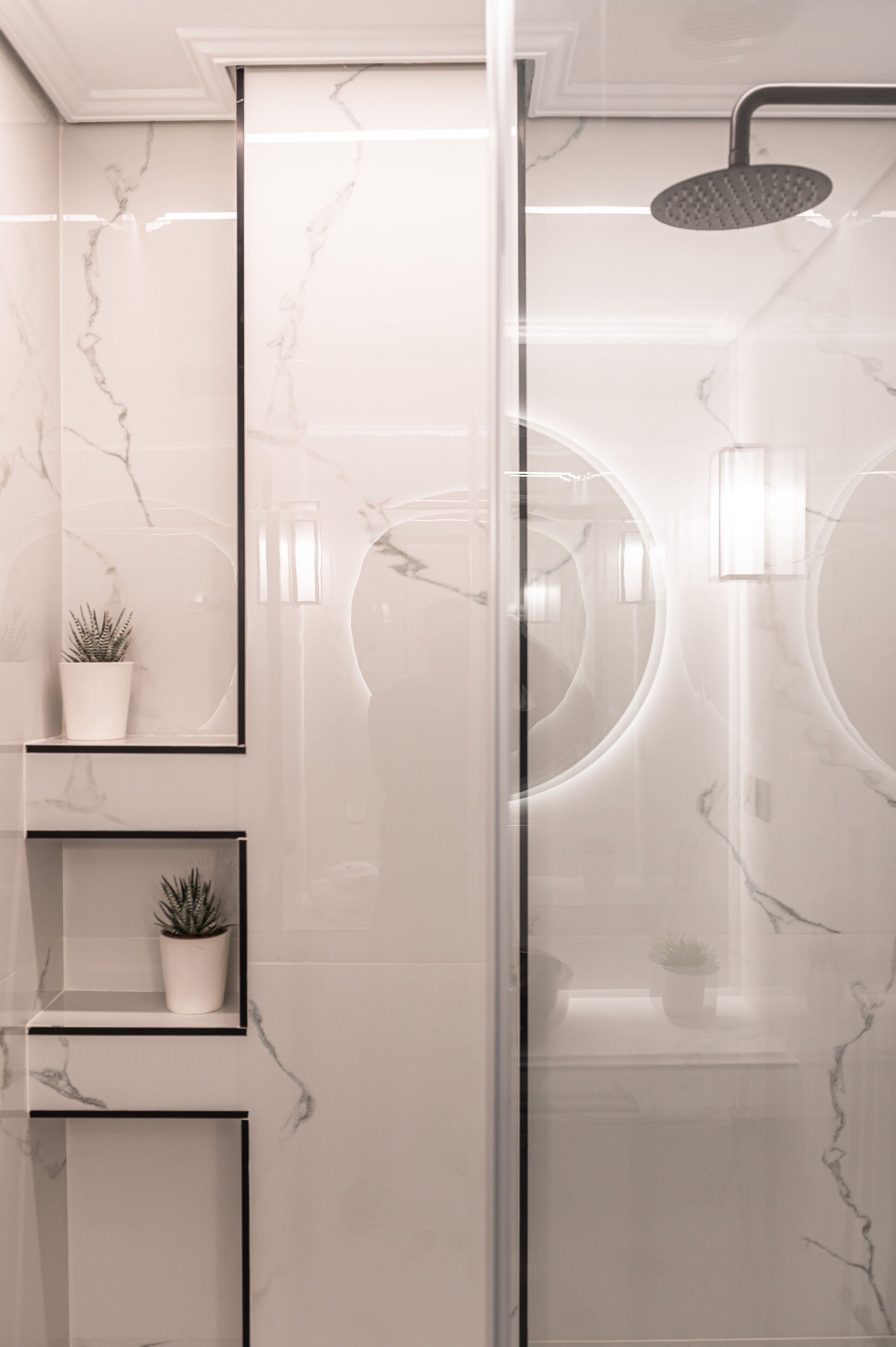 A modern bathroom with marble walls, a glass shower enclosure, and built-in shelves with potted plants.