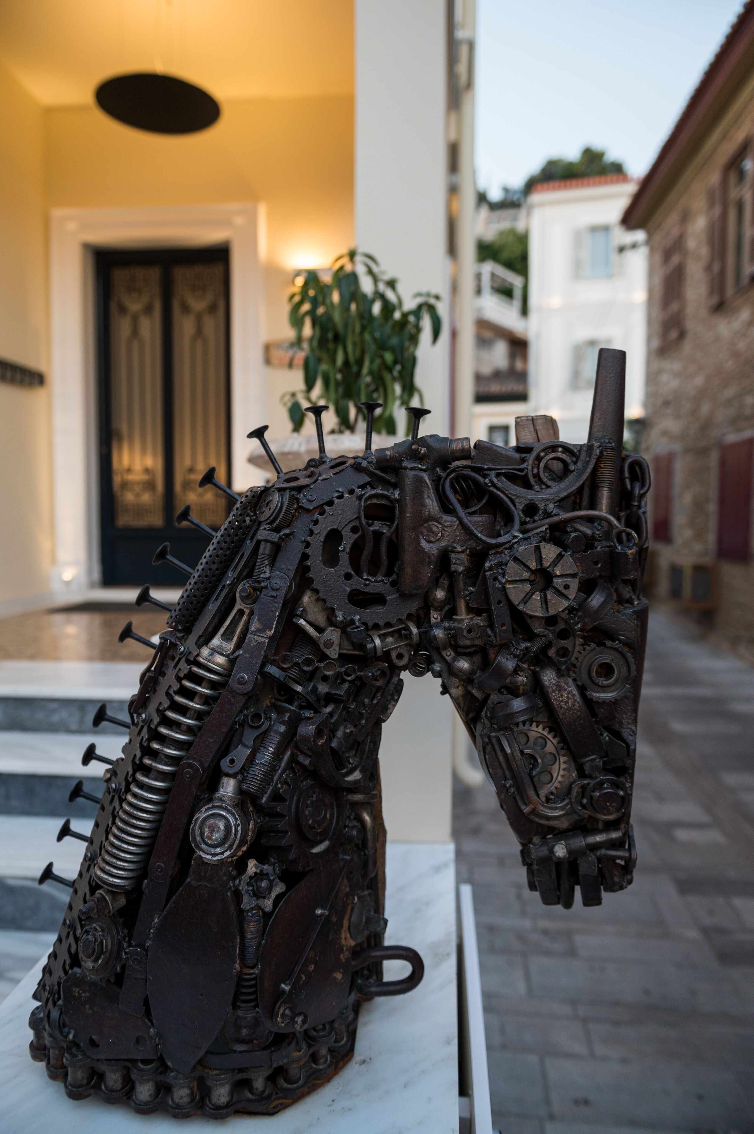 Une sculpture de cheval, réalisée à partir de pièces mécaniques, est placée sur un piédestal blanc dans un espace extérieur.