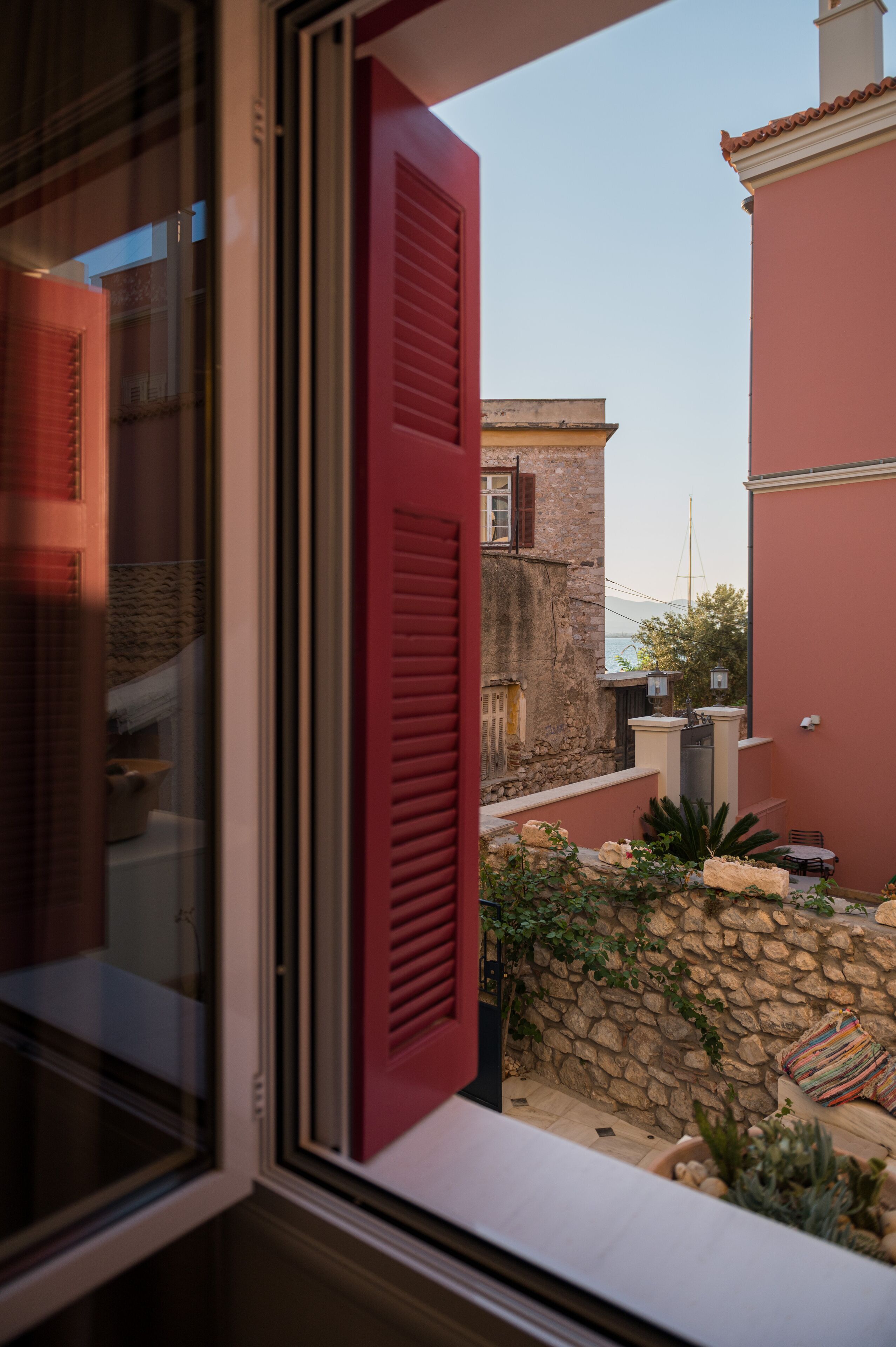 A view through a window with red shutters, revealing a courtyard with a stone wall, a small table, and a chair.