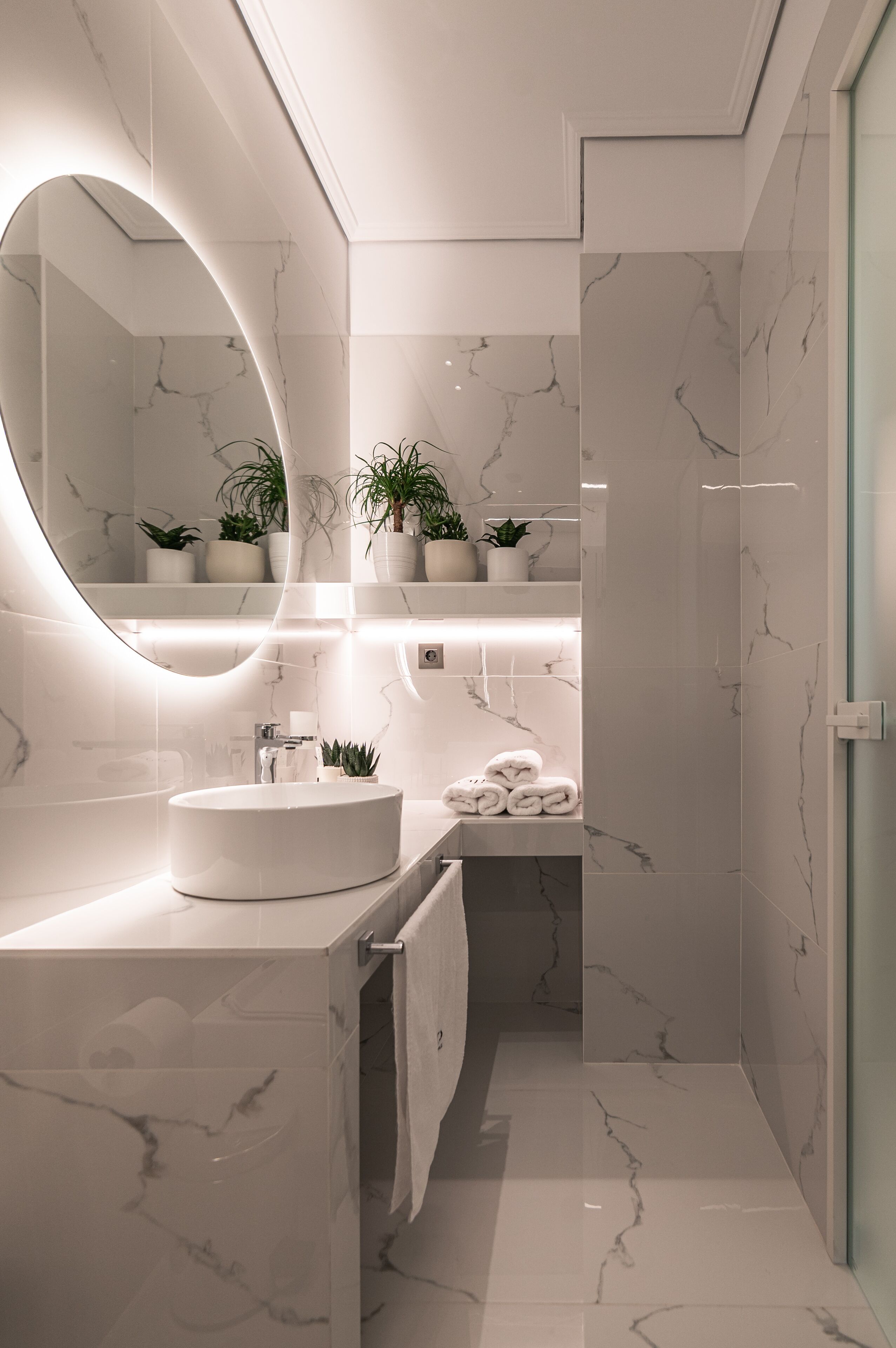 A modern bathroom with a round mirror, white marble tiles, a sink, and potted plants.
