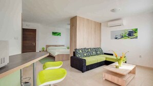 A modern hotel room with a bed, a sofa, a dining table, and a small kitchen area.