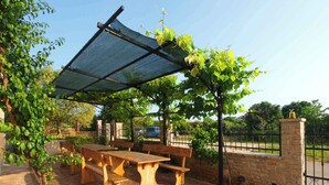 An outdoor patio with a wooden table and benches under a shaded pergola, surrounded by greenery and a stone wall.