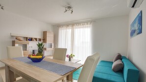 Ein modernes Wohnzimmer mit einem Holzessttisch, einem blaugrünen Sofa und einer kleinen Küchenzeile mit Mikrowelle und einer Ablage für Dekorationen.