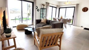 Amplia sala de estar con un sofá grande, una silla de madera y un comedor con mesa y sillas. Amplias ventanas permiten la entrada de luz natural y ofrecen vistas al exterior.