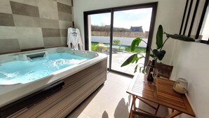 Un baño moderno con una bañera grande, una mesa de madera con una botella y un vaso, y vistas al exterior a través de un ventanal corredero.