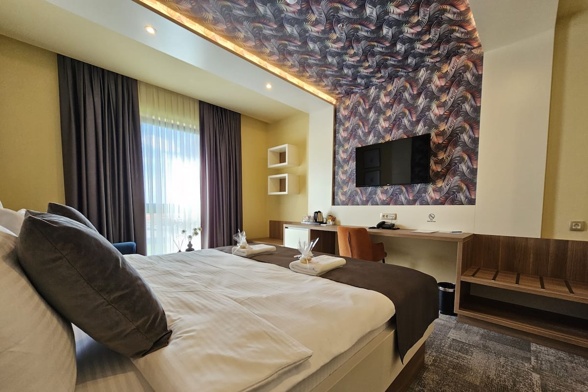 Ein Hotelzimmer mit einem großen Bett, einem Schreibtisch, einem Stuhl, einem Flachbildfernseher an einer Wand mit Blättermotiv und einem Vorhangfenster.