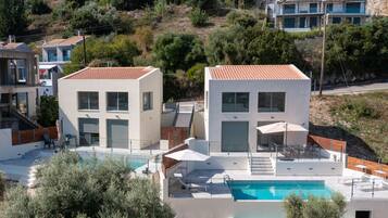 A modern two-story building with a swimming pool, outdoor seating, and a view of surrounding houses.