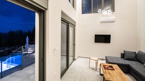 A modern living room with a grey sofa, a wooden coffee table, and a glass door leading to a balcony with a pool. - Elthea Villa 2 in Apolpena, Lefkada (Lefkada)