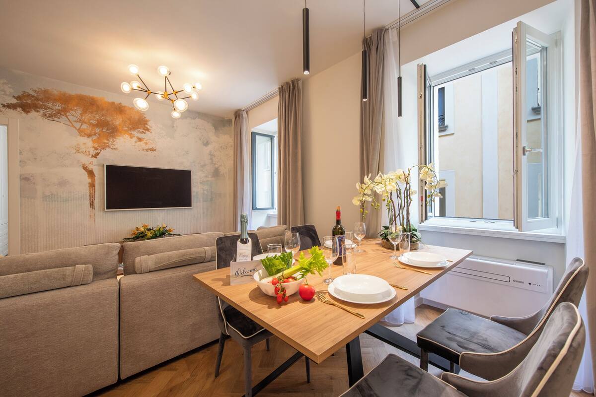 A modern living room with a sofa, a dining table set with plates, glasses, and a bottle. There's a TV mounted on the wall, a chandelier, and a window with curtains.