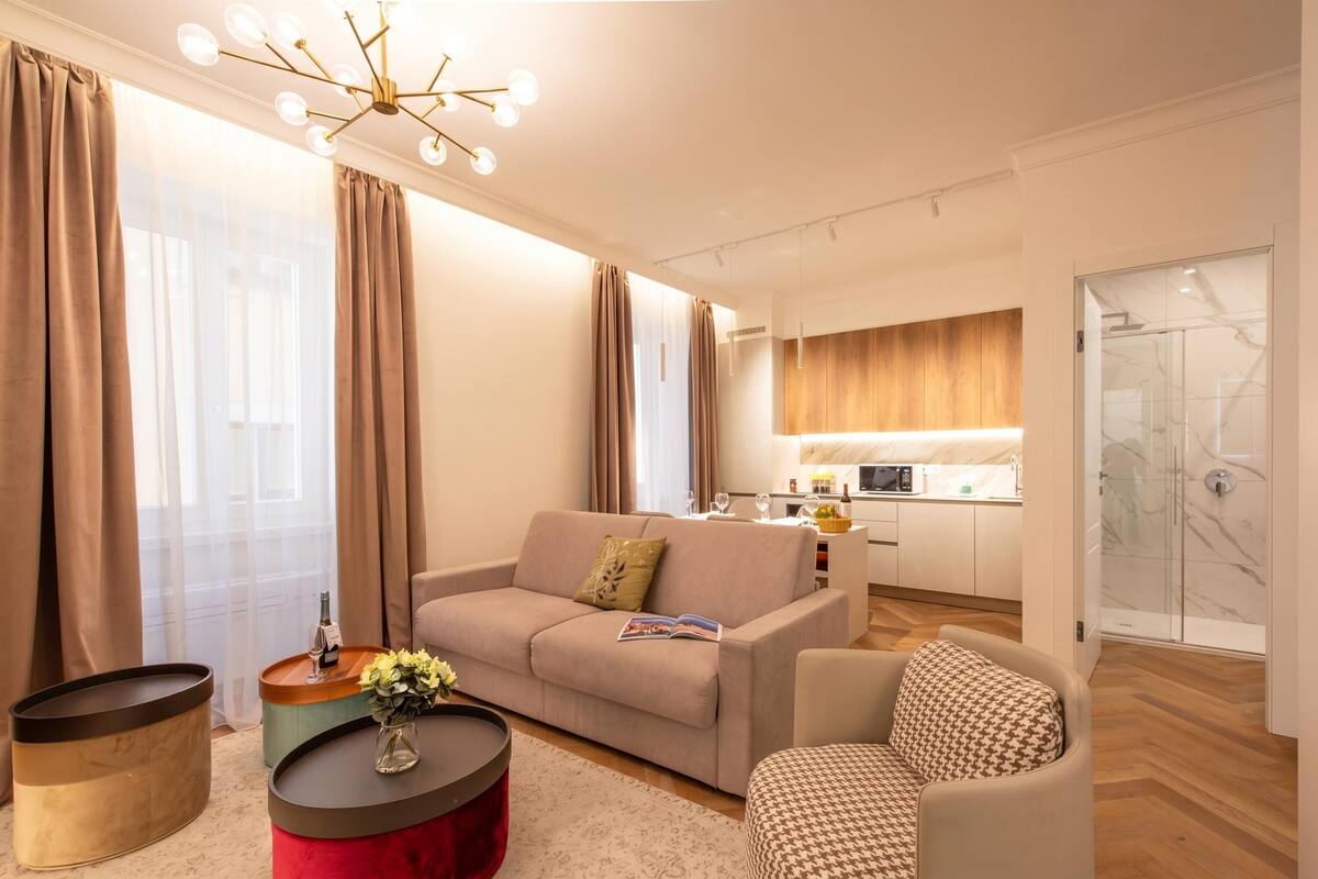 A modern living room with a sofa, armchair, and coffee table, a kitchen in the background, and a glass shower area.