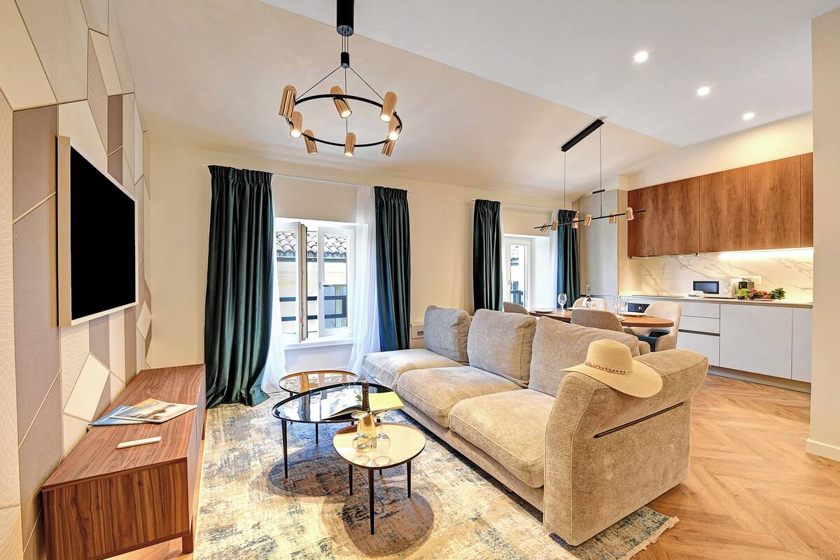 A modern living room with a sofa, coffee table, and TV. There is a kitchen area with cabinets and a dining table.