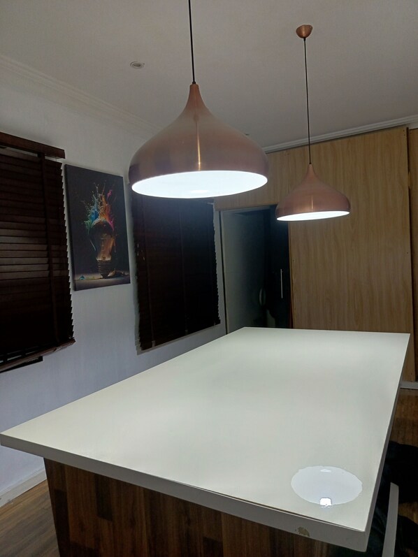 Une cuisine moderne avec un plan de travail blanc, deux suspensions lumineuses, un meuble en bois et une porte dont l’intérieur est de couleur foncée.