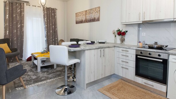 A modern kitchen with white cabinets, a marble countertop, a built-in oven, and a dining area with a bar stool and a table. - Ano Kipseli Athens Gem 1 Bd Apt for 4 (Athens)