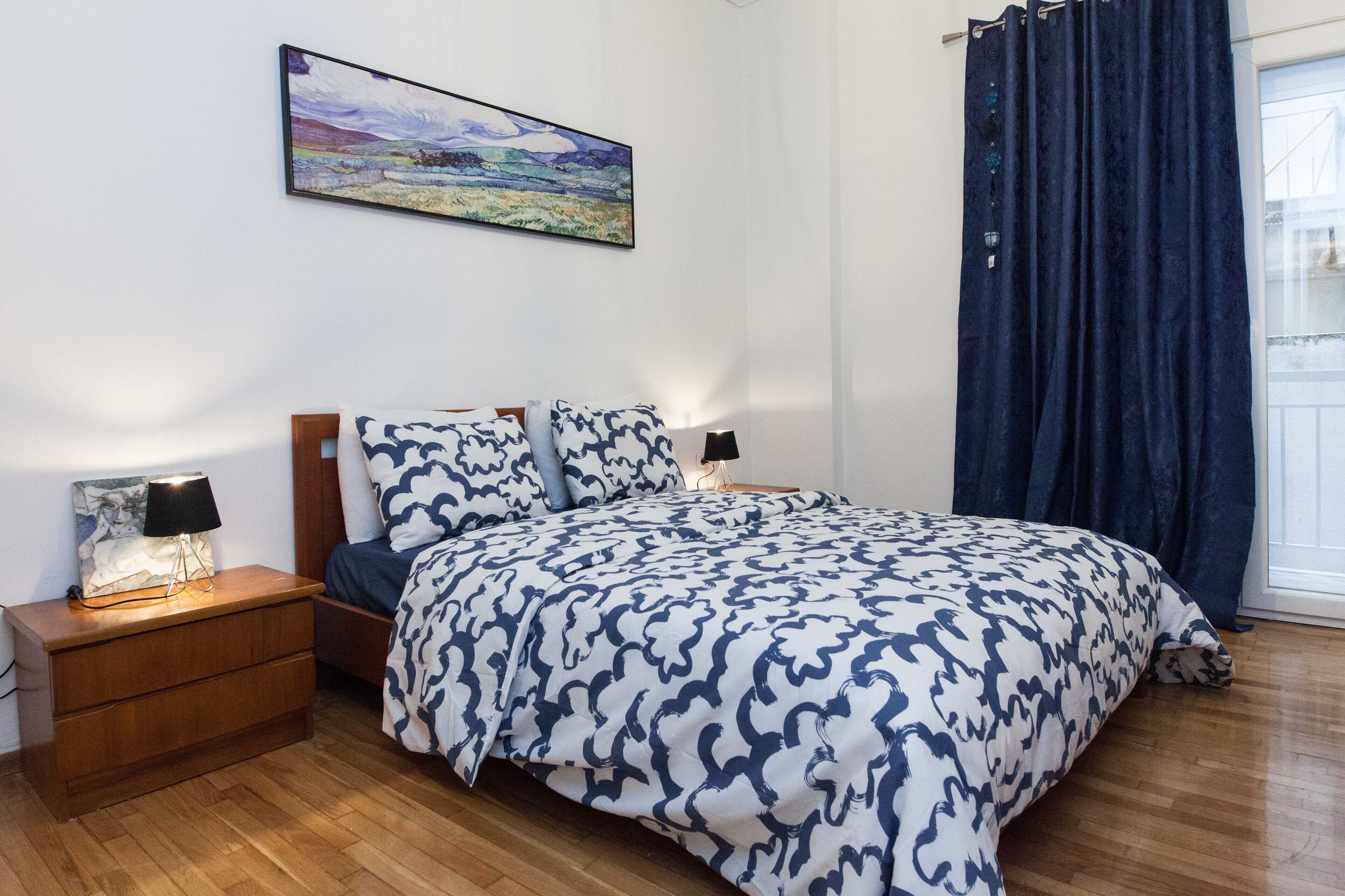 A bedroom with a bed, bedside tables, a lamp, a painting, and a window with curtains.