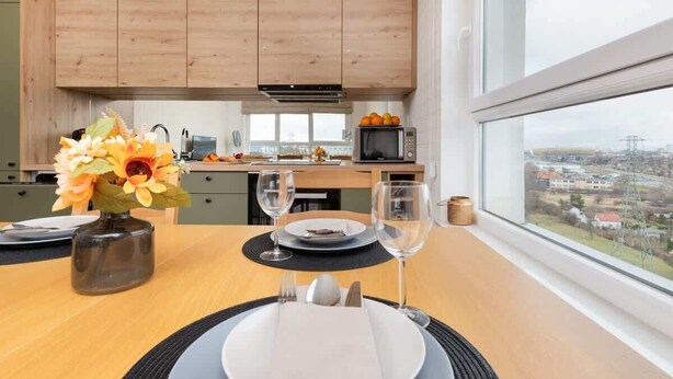 A modern kitchen with wooden cabinets, a dining table set for two with plates, glasses, and a vase of flowers.