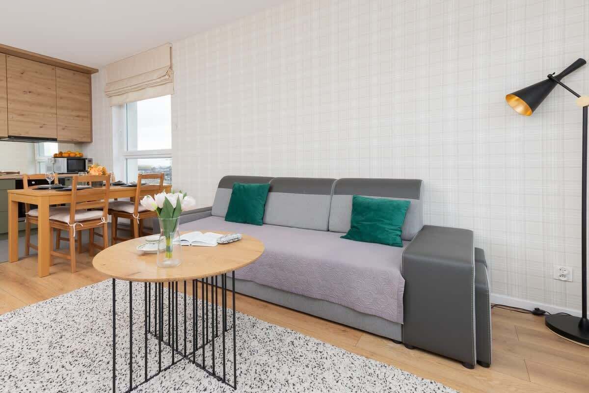 Uma sala de estar moderna com um sofá cinza, uma mesa de centro redonda de madeira, e uma área de jantar com cadeiras de madeira, com uma pequena cozinha ao fundo.