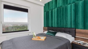 A modern bedroom with a large bed, a green upholstered headboard, a window with a city view, and a bedside table with a tray of breakfast.