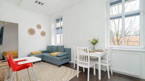 A modern living room with a blue sofa, a white dining table with chairs, a red high chair, and a patterned rug.