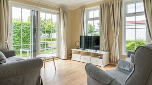 A modern living room with a flat-screen TV, grey sofas, and large windows with curtains.