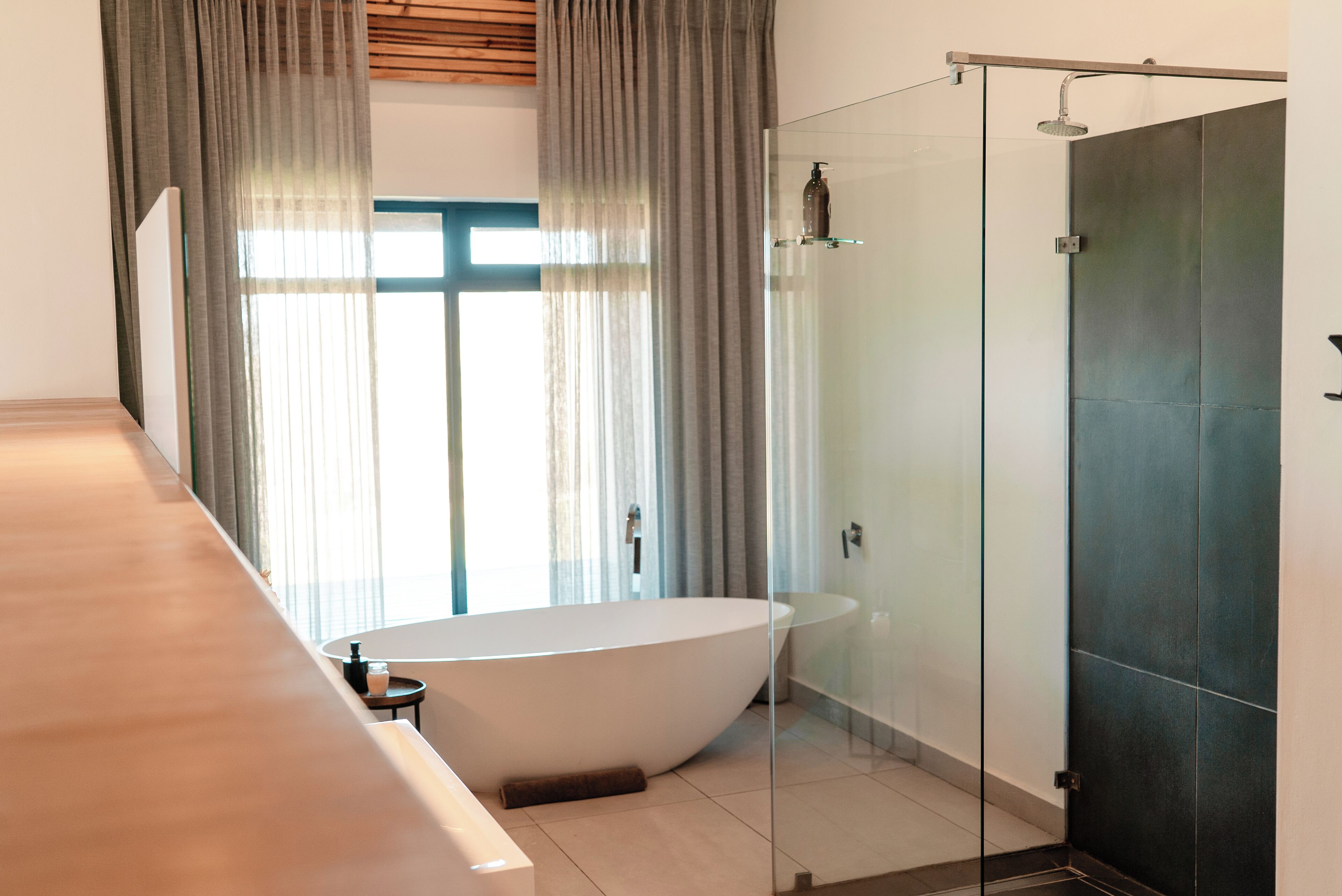 A modern bathroom with a freestanding bathtub, a glass-enclosed shower, and a large window with curtains.