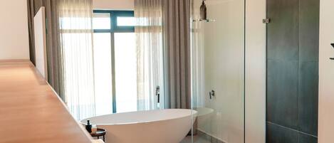 A modern bathroom with a freestanding bathtub, a glass-enclosed shower, and a large window with curtains.