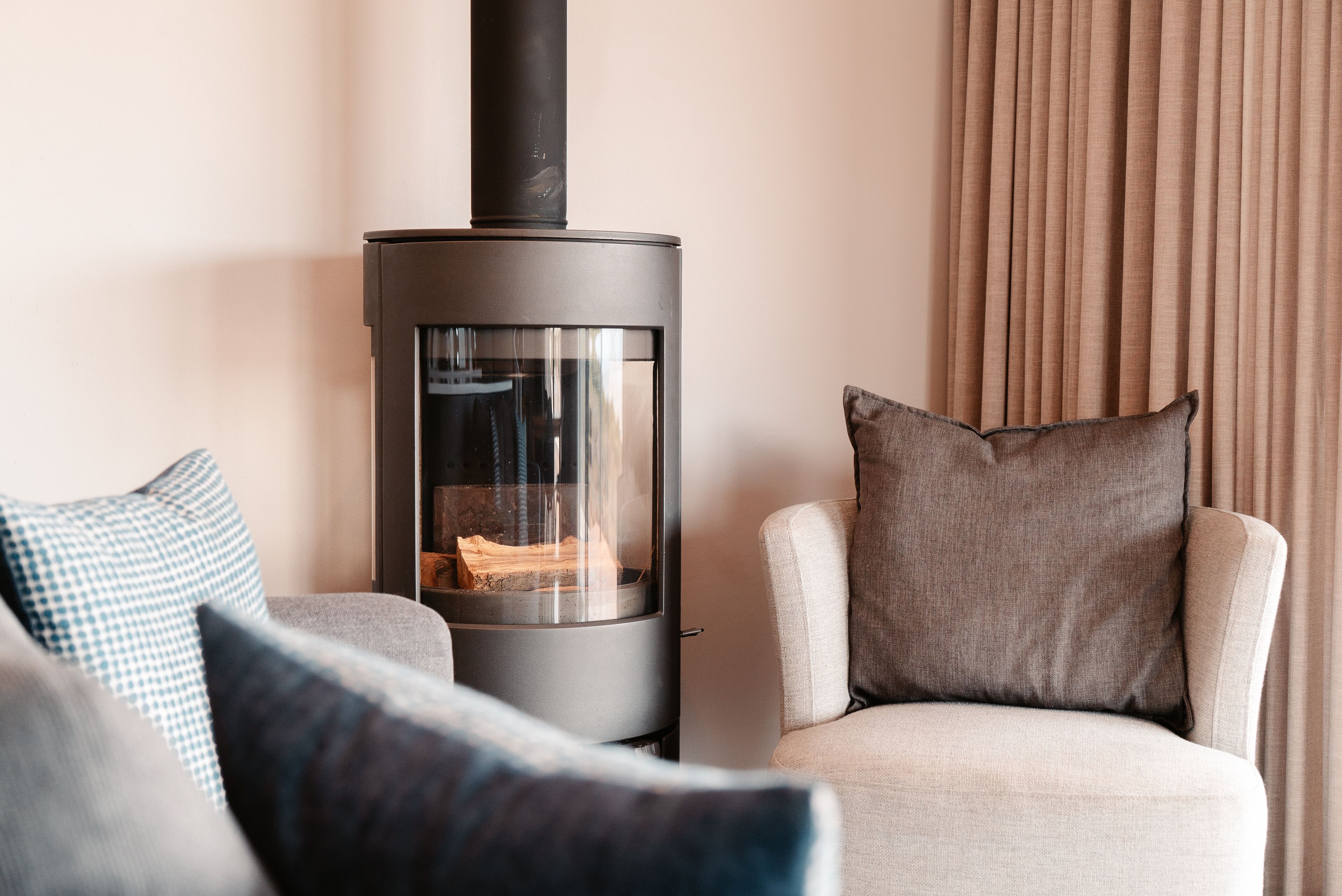 A modern living room with a wood-burning stove, a beige armchair, and patterned cushions.