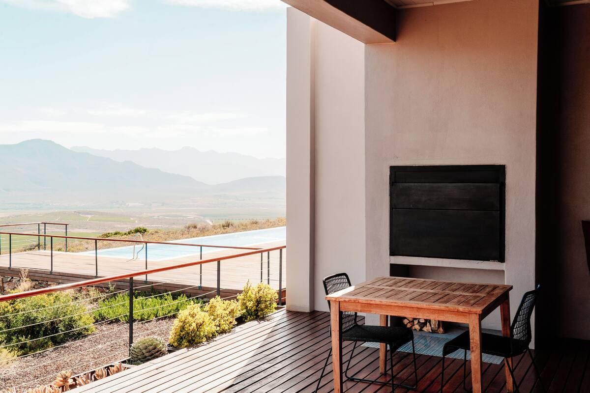 A wooden deck with a table and chairs, overlooking a pool and mountains.