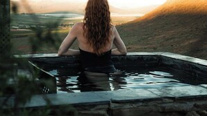 Una persona sentada en una bañera de piedra con vistas a un valle y montañas al fondo.